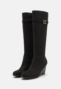 Bottes noires en suède, hauteur genou, avec un talon compensé et une sangle latérale ornée d'une boucle dorée. Texture lisse et design à bout rond.