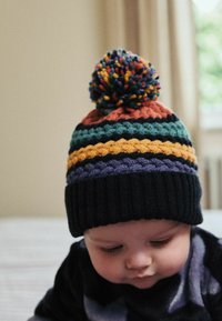 Die gestrickte Mütze hat mehrfarbige Streifen (schwarz, teal, orange, lila) und einen großen, mehrfarbigen Pompon oben. Rippenkante für guten Sitz.