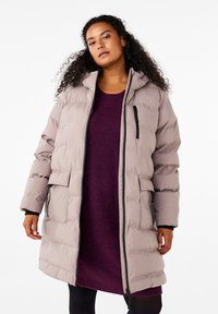 Beige puffjärnjacka med huva, sidofickor och en dragkedjeficka. Bärs över en lila tröjklänning. Mjuk textur, avslappnad passform.