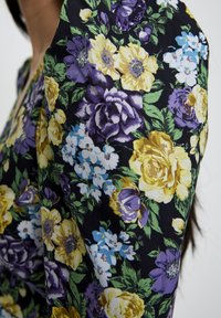 Tessuto di abito floreale con grandi rose gialle e viola, fiori blu e foglie verdi su uno sfondo nero; texture liscia.