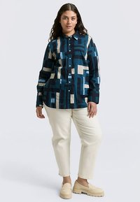 Camicia con bottoni caratterizzata da un motivo geometrico nei colori verde petrolio intenso, blu notte e crema, abbinata a pantaloni chiari e calzature neutre.