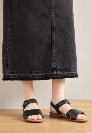 Czarne skórzane sandały z cienkim paskiem, regulowanym zapięciem i wygodną, brązową gumową podeszwą. Noszone z długą dżinsową spódnicą.