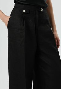 Zwarte linnen broek met een hoge taille, voorzien van twee voorvakken en dubbele zilveren knopen. De stof heeft een gladde, gestructureerde afwerking.