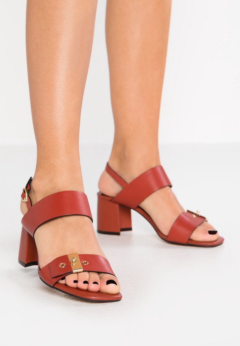 Pieds dans des sandales rouges en cuir à talon bloc avec une boucle dorée et un vernis noir sur les orteils, sur un fond blanc uni.