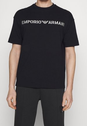 Homme portant un t-shirt noir à manches courtes avec un texte et un logo argentés "EMPORIO ARMANI" sur la poitrine.