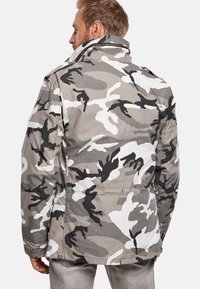 Chaqueta de camuflaje en gris, blanco y negro. Cuenta con un cuello estructurado, múltiples costuras y una textura de tela detallada.