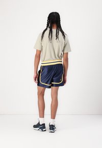 T-shirt à manches courtes, vert olive clair, associé à un short de sport bleu marine avec une ceinture élastique jaune et blanche et des accents sur les côtés.