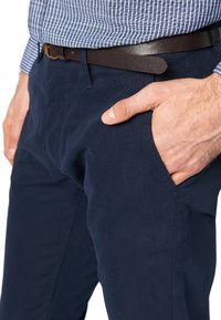 Mężczyzna w granatowych spodniach i brązowym pasku, z jedną ręką opartą w przedniej kieszeni, ubrany w niebieską koszulę w kratę.