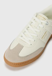 White sneaker with a beige suede toe cap and leather body. Features white laces, an embossed logo, and a brown rubber sole.