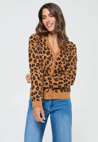Cardigan à motif léopard marron avec des manches longues et un décolleté en V, doté de boutons marron et d'une texture en maille douce. Associé à un jean bleu.