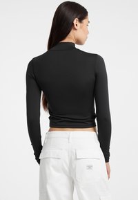 Top corto nero a maniche lunghe con collo alto, realizzato in tessuto elasticizzato. Indossato con pantaloni cargo bianchi con tasche laterali.