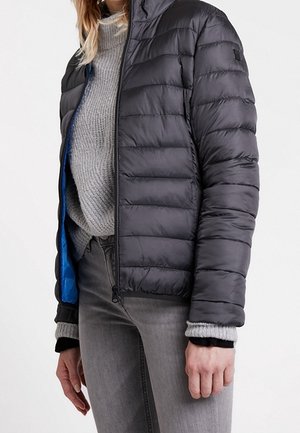 Person wearing a black quilted jacket over a gray knit sweater and gray jeans, with jacket partially unzipped showing blue lining.