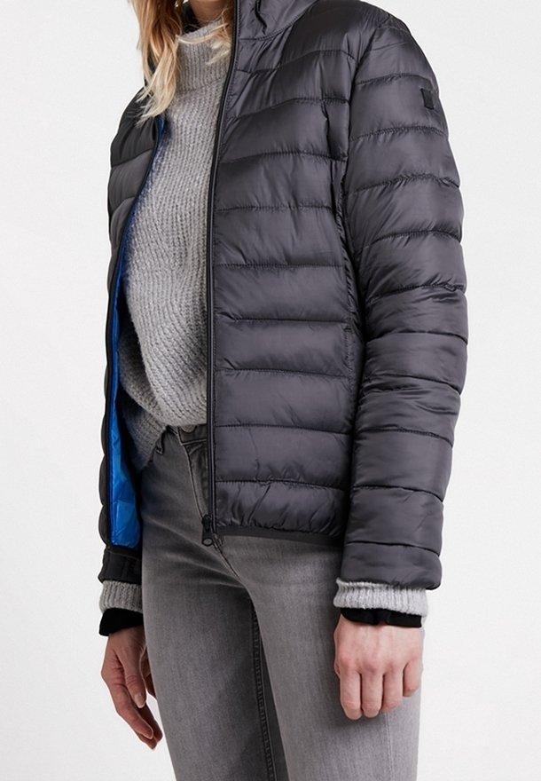 Personne portant une veste matelassée noire par-dessus un pull gris en maille et un jean gris, la veste partiellement ouverte laissant apparaître la doublure bleue.