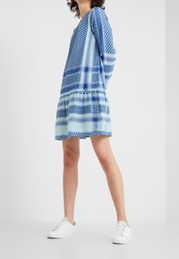 Sininen kuviollinen mekko, jossa on pitkät hihat ja kerroksellinen hame. Valmistettu kevyestä kankaasta, jossa on erilaisia sinisiä sävyjä ja geometrisia kuvioita.