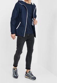 Merijalkoinen tuulitakki valkoisilla yksityiskohdilla, yhdessä tummansinisen t-paidan, tummanharmaiden farkkujen ja harmaankeltaisten lenkkareiden kanssa, jossa on teksturoitu pohja.