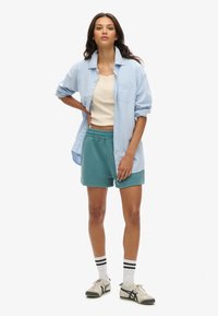 Camisa de botones azul claro sobre una camiseta sin mangas beige, combinada con pantalones cortos verde azulado y calcetas blancas con rayas negras, junto con zapatillas neutrales.