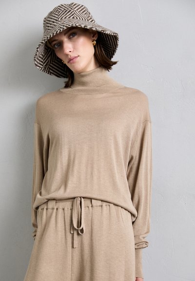 Beige, long-sleeve, high-neck top with loose fit, paired with matching pants that have a drawstring waist. Accented by a striped bucket hat.