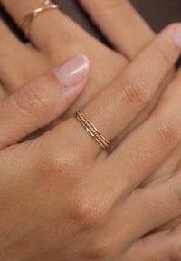 Tre anelli d'oro sovrapposti su una mano, con una texture intrecciata, fasce lisce e una finitura lucida. Pelle chiara e unghie naturali.