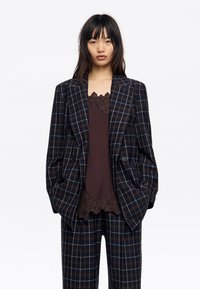 Marynarka w kratę w kolorze ciemnogranatowym, brązowym i białym, z dwoma guzikami, wąskimi klapami i luźnym krojem, noszona na brązowym topie z koronkową lamówką.