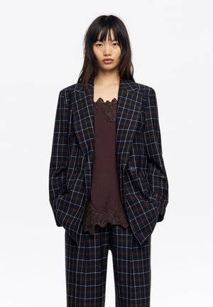Marynarka w kratę w kolorze ciemnogranatowym, brązowym i białym, z dwoma guzikami, wąskimi klapami i luźnym krojem, noszona na brązowym topie z koronkową lamówką.