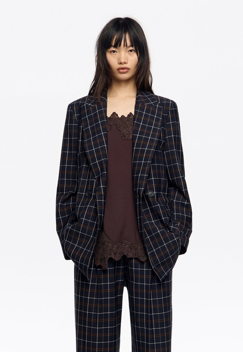 Marynarka w kratę w kolorze ciemnogranatowym, brązowym i białym, z dwoma guzikami, wąskimi klapami i luźnym krojem, noszona na brązowym topie z koronkową lamówką.
