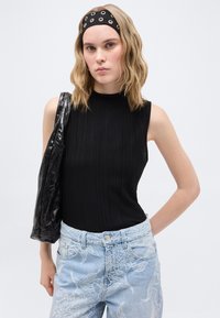 Νεαρή γυναίκα με κυματιστά ξανθά μαλλιά φοράει μαύρο αμάνικο τοπ, μαύρη κορδέλα με τρύπες, ανοιχτό μπλε τζιν με κεντήματα και κρατάει μαύρη τσάντα ώμου.