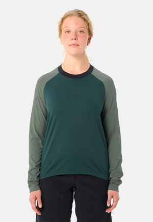 Jonge persoon met krullend blond haar draagt een donker groen-grijs shirt met lange mouwen en een zwarte broek, staande tegen een witte achtergrond.