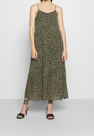 Kvinde iført en grøn blomsterprintet ærmeløs midi-kjole med spaghetti stropper og sorte high-heel sandaler med stropper, stående på en grå gulv.