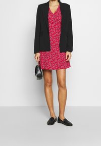 Schwarzer maßgeschneiderter Blazer über einem roten, blumengemusterten Slipkleid. Schwarze flache Schuhe und eine kleine schwarze Handtasche. Glattes Material, knielanges Kleid.