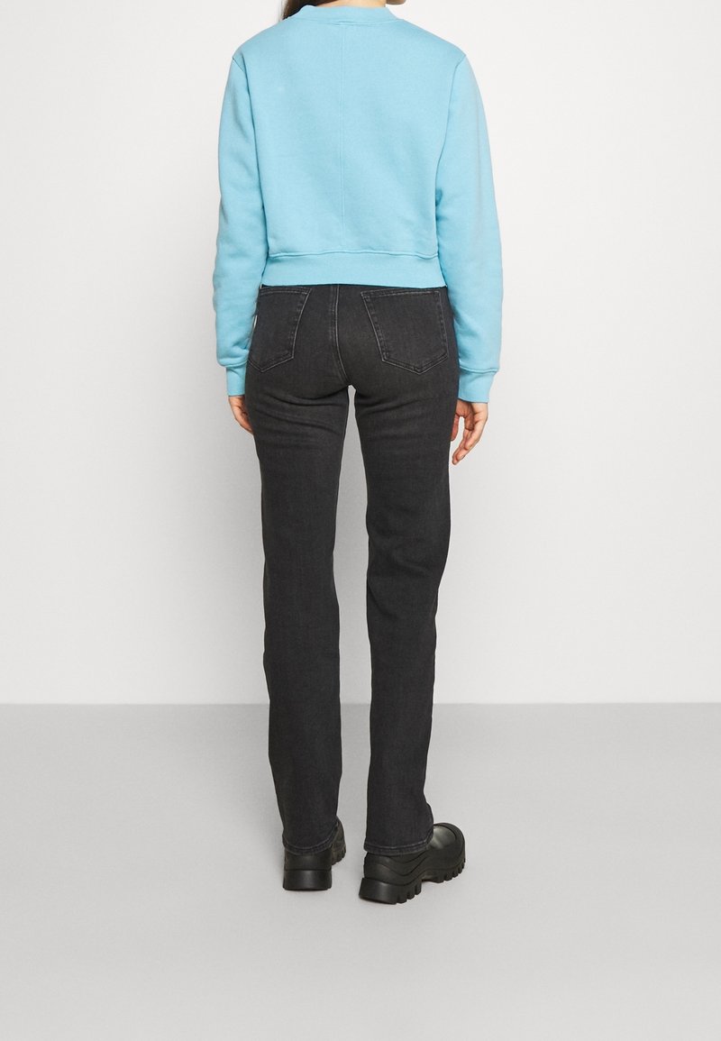 Personne portant un sweat-shirt bleu clair, un jean noir et des chaussures noires épaisses, debout de dos sur un fond blanc uni.