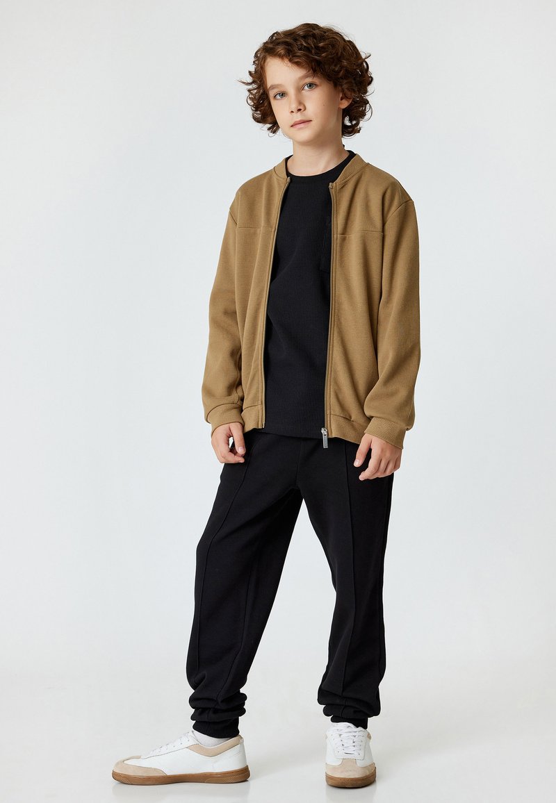 Brun zip-up jakke over en sort skjorte, parret med sorte joggere og hvide sneakers med beige detaljer. Glatte teksturer, afslappet pasform.