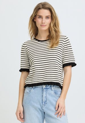 Donna con capelli mossi sciolti che indossa un maglione a maniche corte a righe bianche e nere e jeans azzurri a vita alta su uno sfondo semplice.