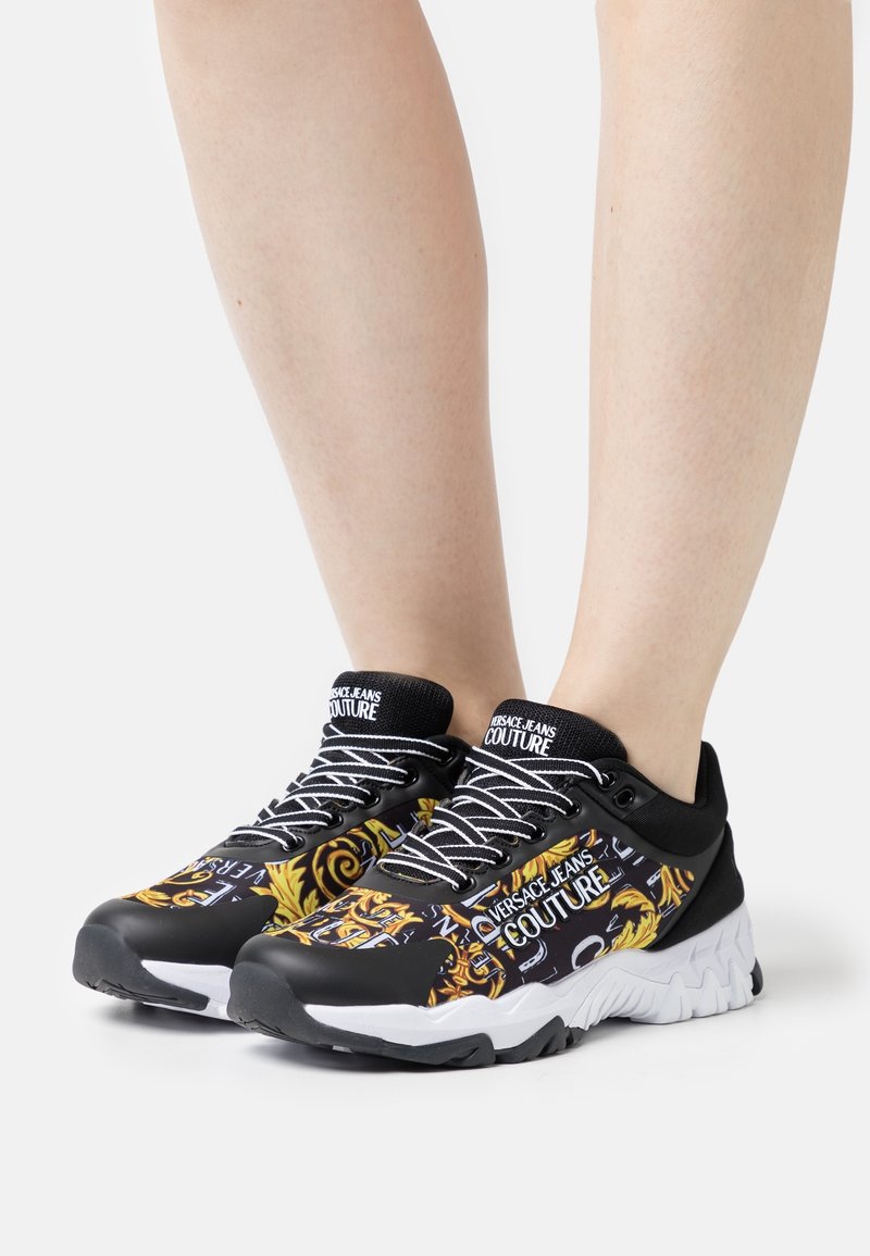 Jambes portant des baskets Versace Jeans Couture à motifs noirs, blancs et dorés avec des lacets blancs sur un fond blanc.