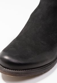 Bota de cuero negro hasta el tobillo con un acabado liso y punta redonda. Presenta una suela resistente con un sutil detalle de costura alrededor del borde.