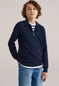 Jeune personne aux cheveux bouclés portant un pull zippé bleu marine texturé sur une chemise à rayures et un jean bleu clair, debout avec une main derrière le dos.