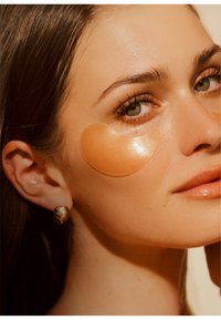 Patch per gli occhi in idrogel di un leggero colore marrone traslucido, applicati sotto a occhi verdi con ciglia definite, pelle con un finish luminoso e orecchini d'oro.