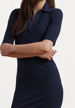 Femme portant une robe côtelée bleu marine à manches courtes et col, bras croisés à la taille, sur un fond clair uni.