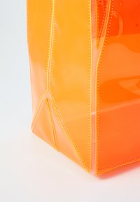 Transparent orange tote bag med en blank overflate, gule sømmer, og et geometrisk bunndesign. Kantene er avrundede, noe som fremhever formen.