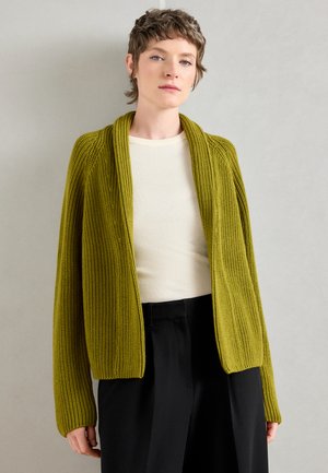 Cardigan vert olive avec texture côtelée, ouvert à l'avant et manches longues, associé à un haut fitted beige clair et un pantalon noir.