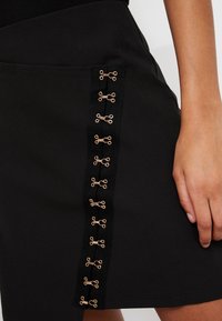 Gonna nera con pannello laterale accentuato da hardware dorato. Tessuto liscio, design aderente e silhouette elegante.