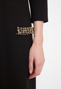 Gros plan d'une main à la peau claire reposant à côté d'un vêtement noir avec un détail rectangulaire en métal doré clouté au niveau de la hanche.