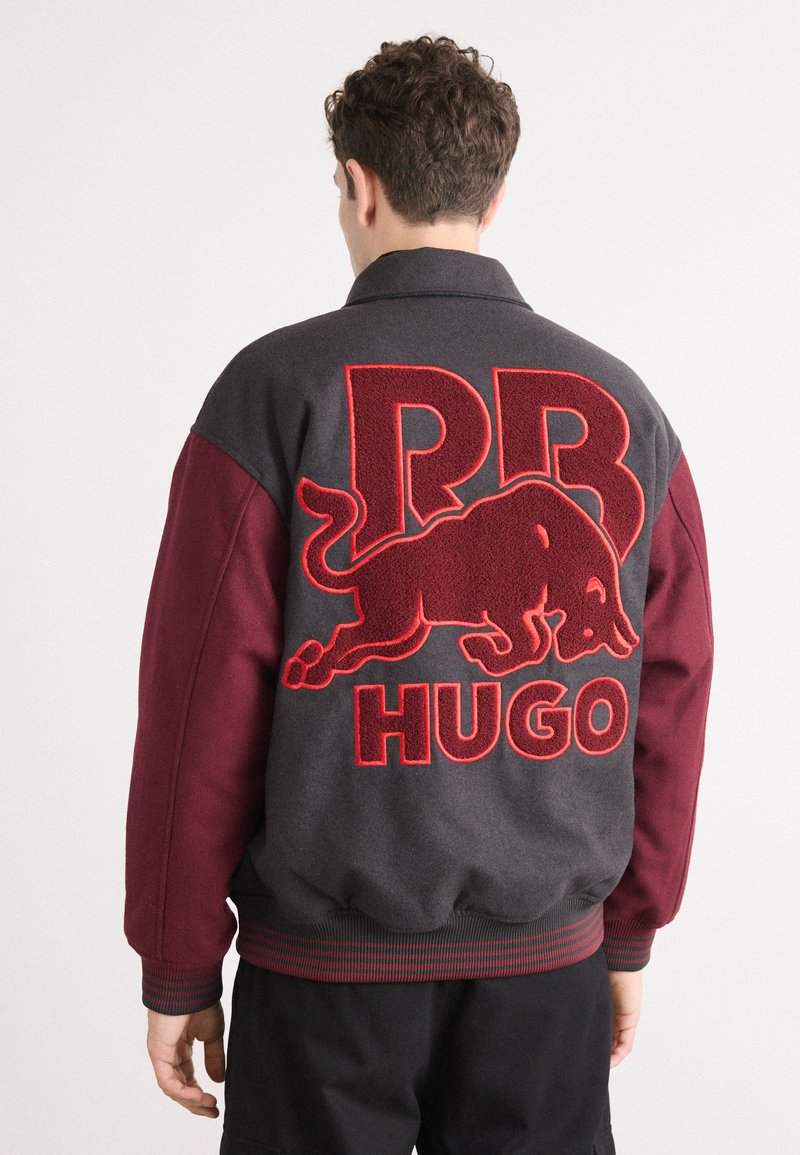 Grå og burgunder bomberjakke med stort rødt broderet 'RB HUGO' logo og tyr design på ryggen, med ribbetrøje og søm.