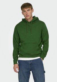 Felpa verde con cappuccio, tasca a marsupio, cordini e polsini a coste, indossata sopra una maglietta bianca e abbinata a jeans blu.