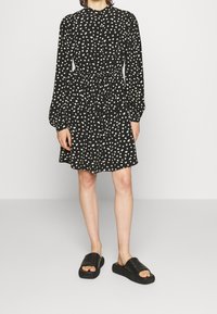 Vestido negro con estampado floral blanco, mangas largas, cintura ajustada y falda ligeramente acampanada. Combinado con sandalias negras de tipo slide.