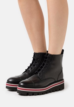 Bottines en cuir noir à lacets avec semelles compensées épaisses à rayures noires, blanches et rouges, portées sur des jambes nues.