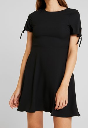 Kobieta ubrana w krótką czarną sukienkę z krótkimi rękawami związanymi w kokardki, stoi na jednolitym jasnoszarym tle.