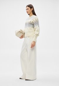 Mulher com camisola creme estampada e calças brancas de pernas largas, segurando uma carteira dobrada bege contra um fundo simples.