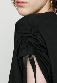 Lähikuva henkilön olkapäästä ja kaulasta, joka on pukeutunut mustaan vaatteeseen, jossa on rypytetyt hihat kiinni mustilla nauhoilla.