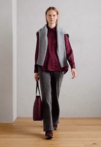 Gilet gris sur une chemise bordeaux à boutons, associé à un pantalon évasé gris foncé et un sac à main bordeaux. Textures lisses, arrière-plan neutre.