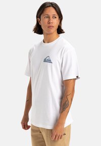 Camiseta blanca de algodón con cuello redondo y mangas cortas, que presenta un gráfico de olas azul y verde en el pecho. Etiqueta en la manga.